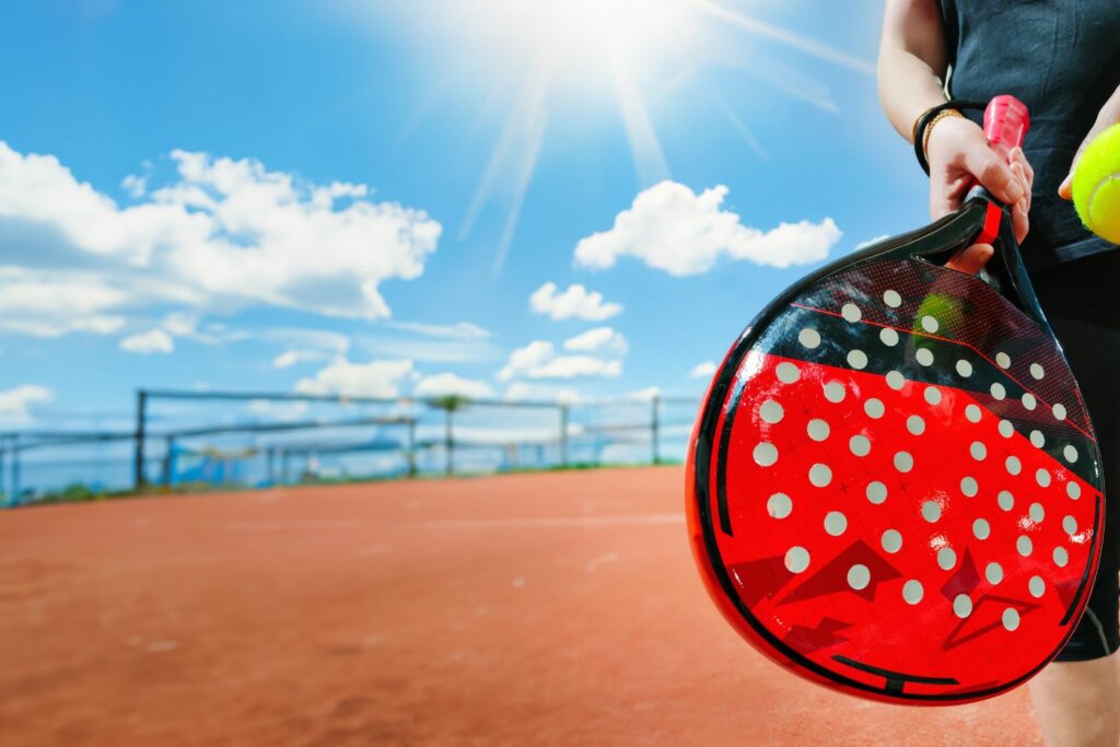 court extérieur de Padel sous le soleil en arrière plan et en premier plan une raquette en larme noir et rouge tenue par une main