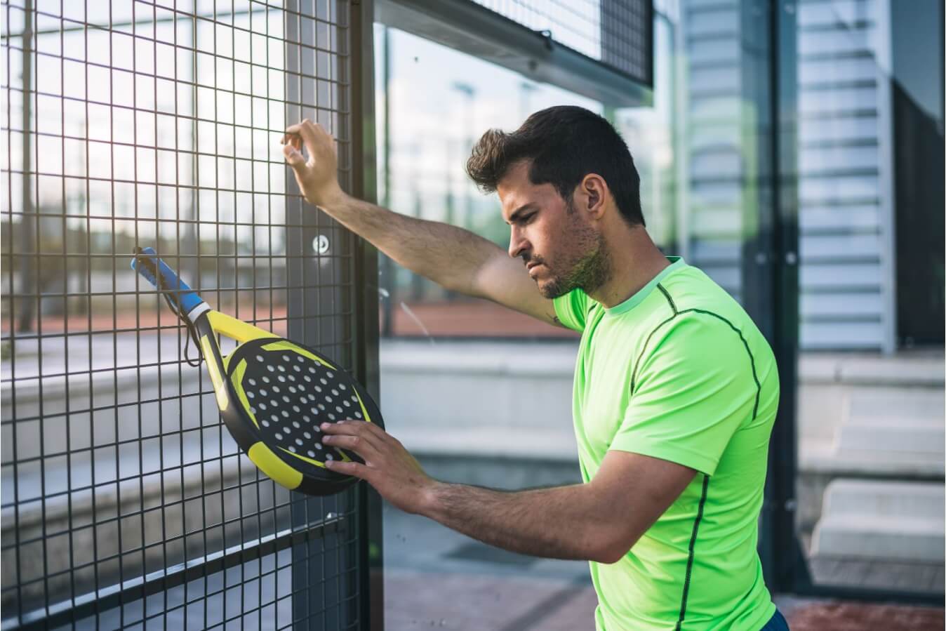 un joueur de Padel qui s'appuie sur un grillage de terrain en fixant sa raquette de Padel