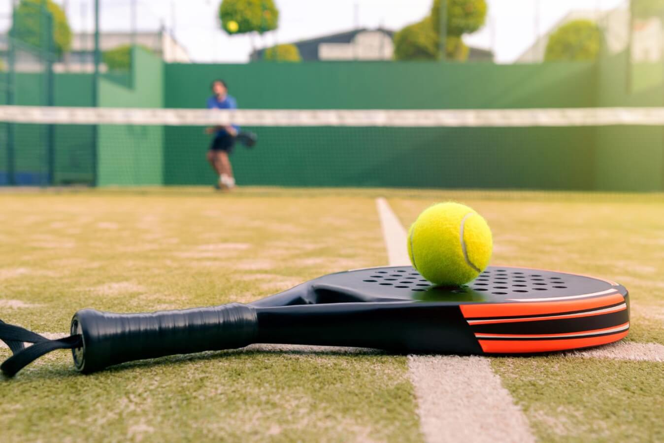 au premier plan, une raquette de Padel noire avec une balle au-dessus, le tout posé sur le sol d'un terrain et en arrière plan, un joueur