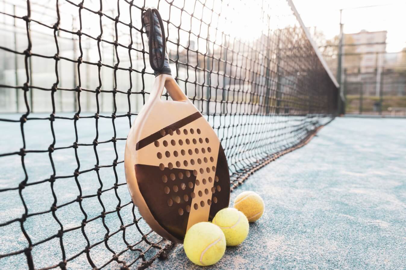 un court de Padel sous le soleil et une raquette de couleur beige et marron adossée au filet et 3 balles jaunes devant