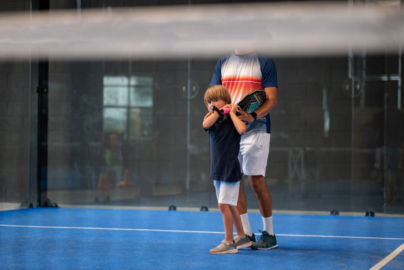 un enfant tenant une raquette et près d'un coach sur un terrain de Padel bleu