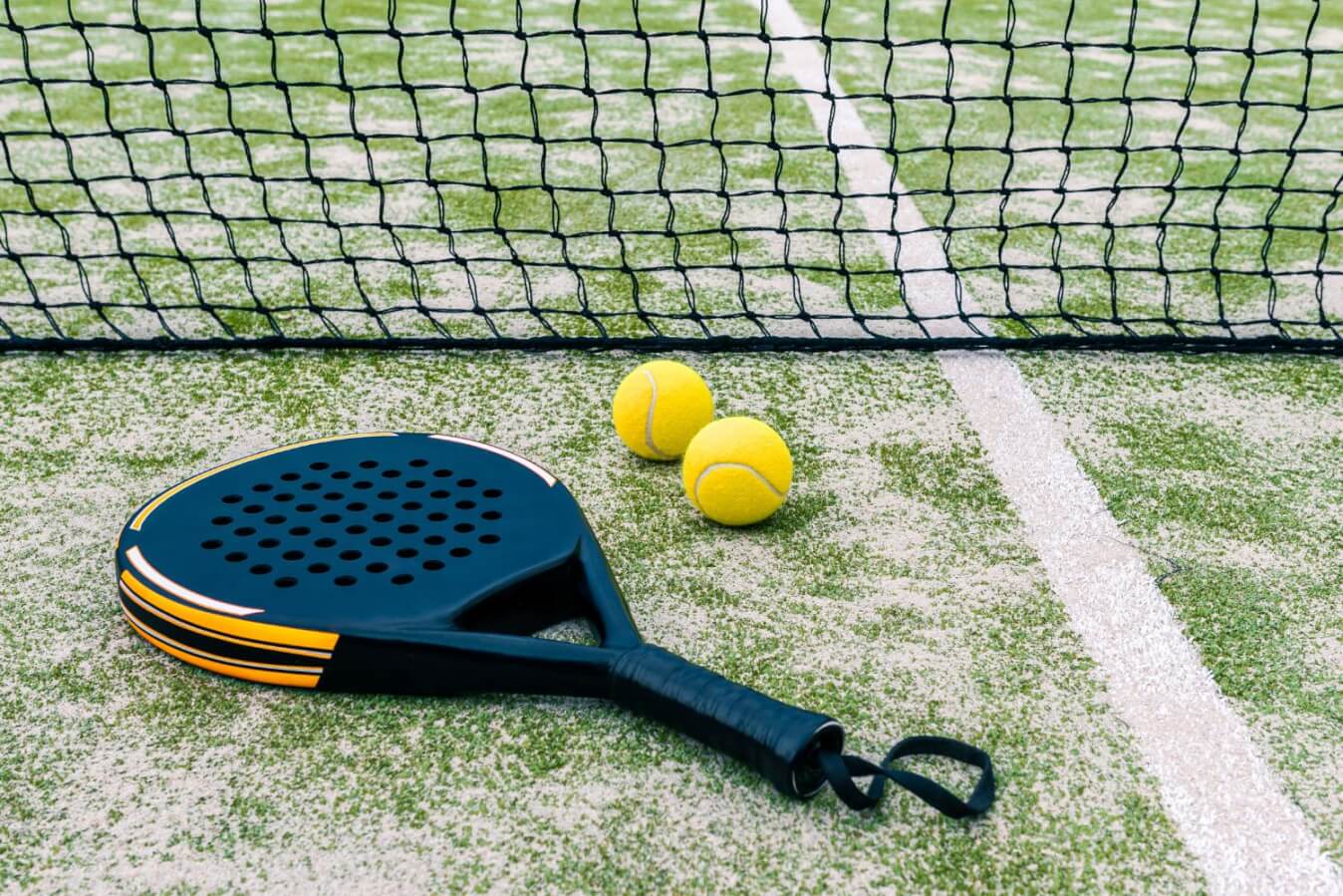 une raquette de Padel pour enfants de couleur noir et orange avec 2 balles jaunes, tous posés sur un sol en gazon, près du filet