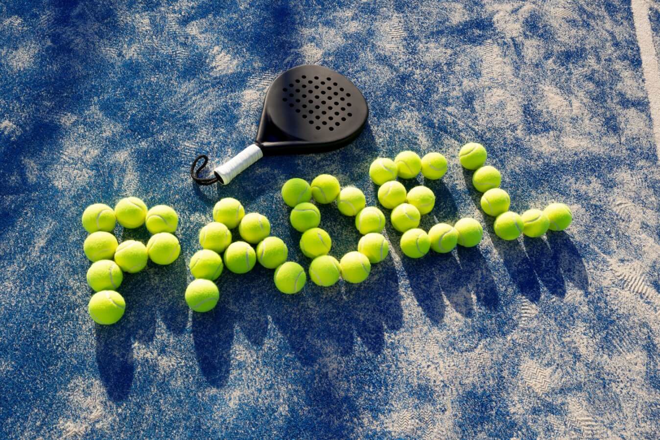 une raquette de Padel enfant noire et des balles qui écrivent Padel posés sur le sol d'un terrain de Padel