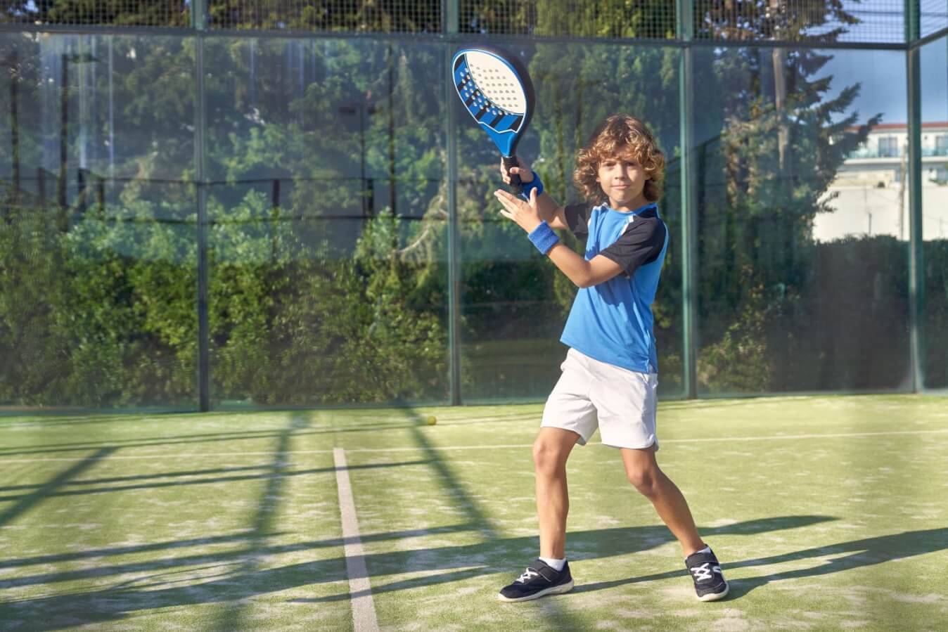 un petit garçon portant un short blanc et un t-shirt bleu tient une raquette de Padel sur un terrain en gazon