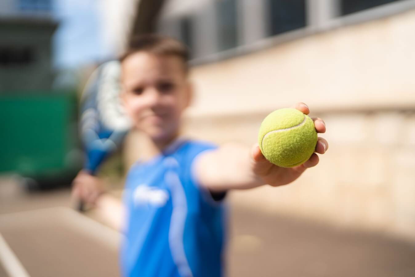 une balle jaune de Padel en premier plan et en flou un petit garçon avec raquette à la main et la balle jaune