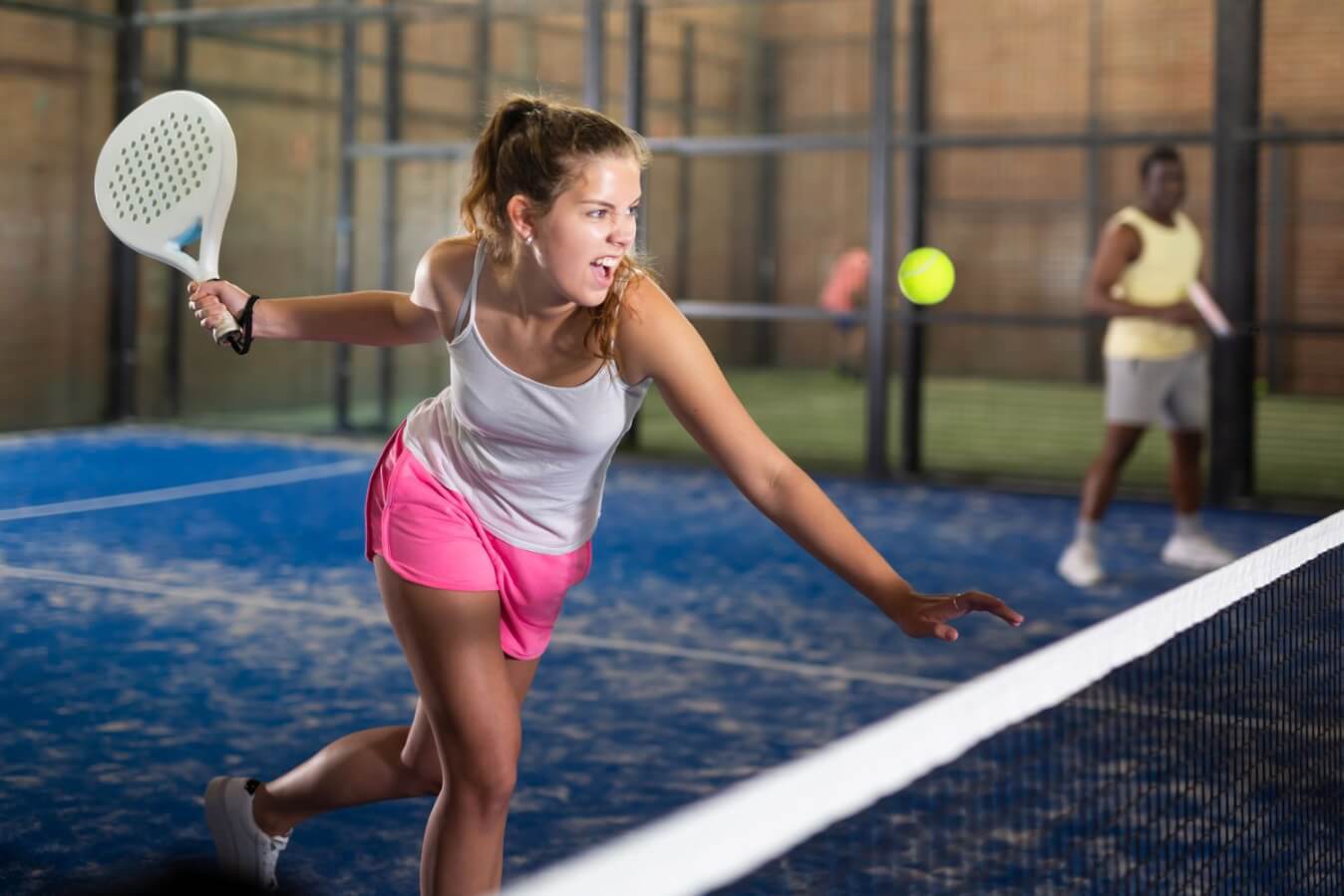 une joueuse de Padel avec raquette blanche à la main prête à frapper la balle jaune devant le filet