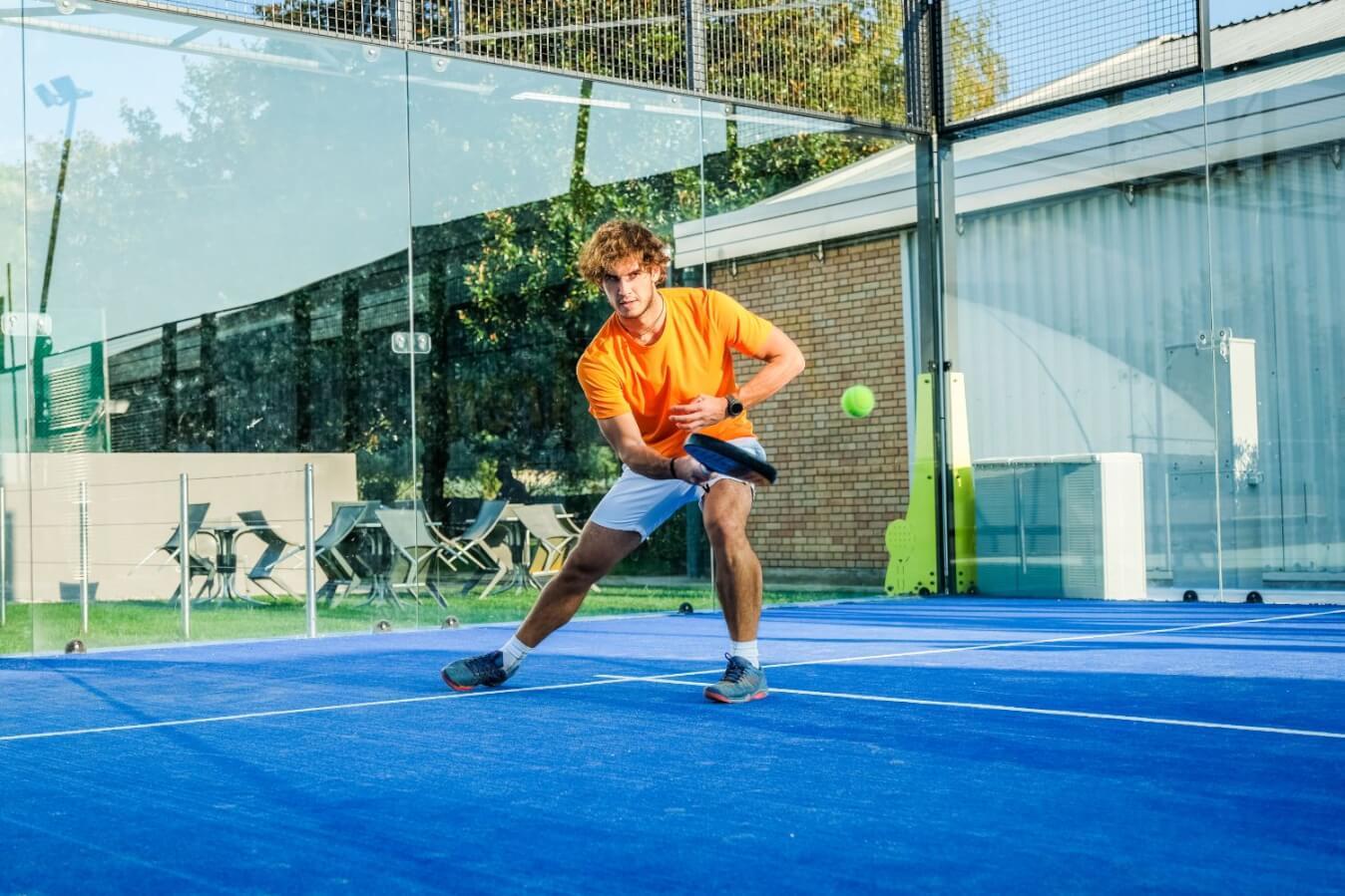 un joueur de Padel en t-shirt orange et short blanc sur un terrain, raquette à la main