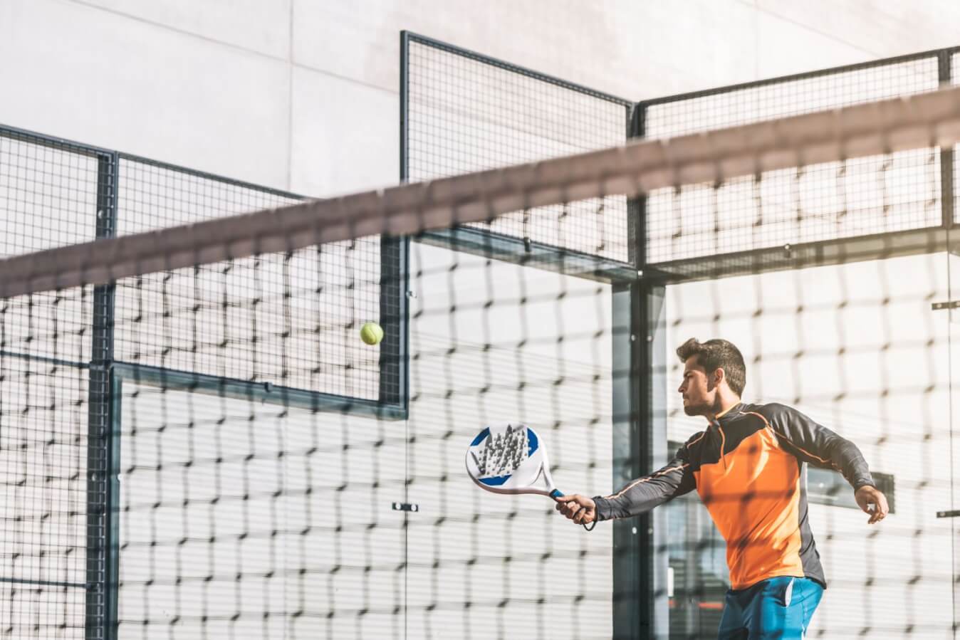 un joueur de Padel en tenue orange et bleu, raquette à la main prêt à frapper la balle