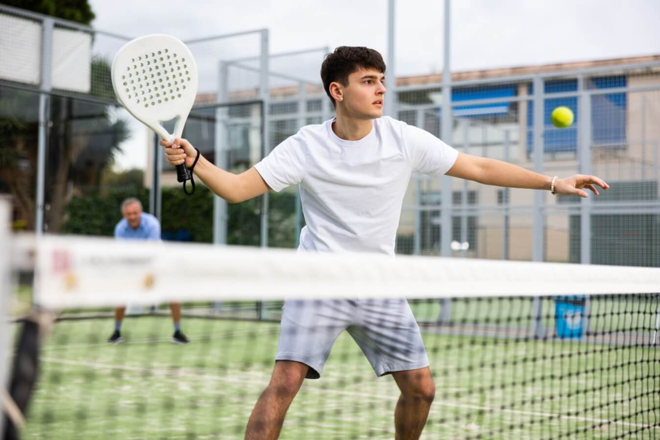 jeune joueur de Padel tout en blanc avec raquette blanc sur un terrain en gazon prêt à frapper la balle jaune près du filet