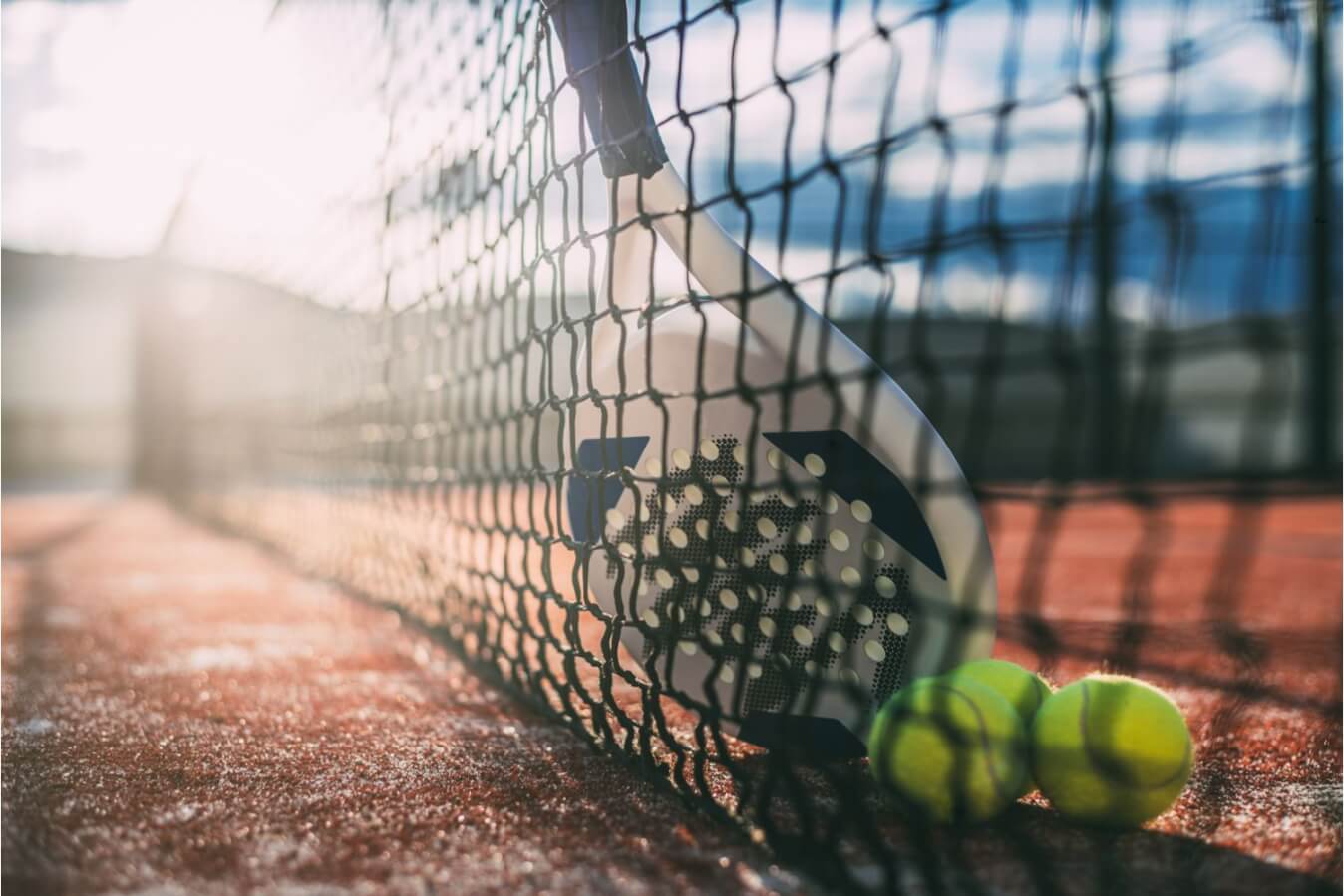 une raquette et 3 balles adossées au filet d'un terrain de Padel sous le soleil