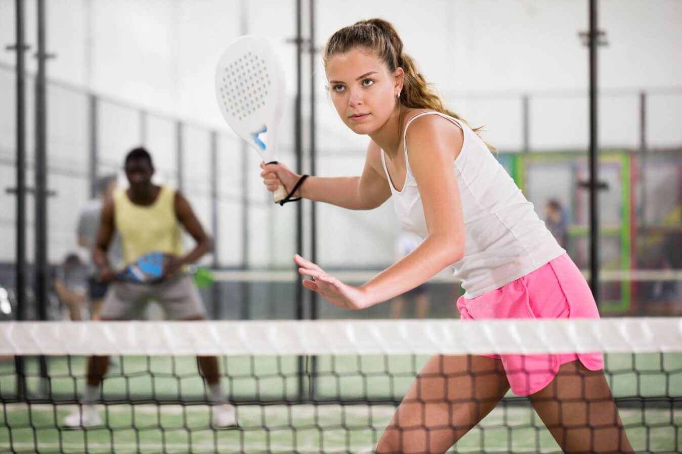 joueuse de Padel avec raquette blanche à la main, portant un short rose et un débardeur blanc, près du filet du terrain de Padel