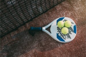 raquette de Padel de couleur blanc et bleu avec manche noir posée sur un terrain près du filet avec 3 balles jaunes sur le dessus