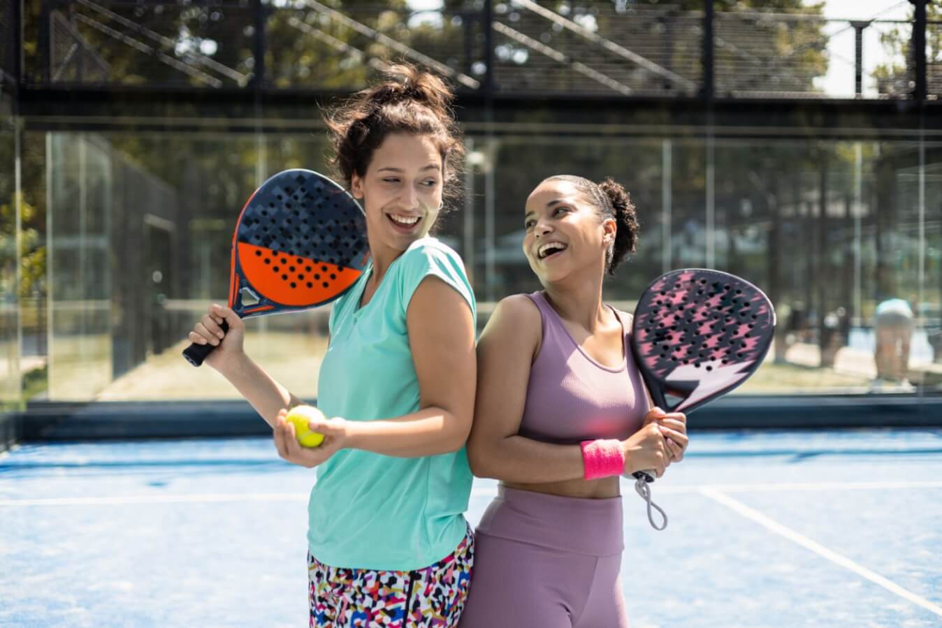 2 jeunes filles se tournant le dos et riant et portant chacune une raquette de Padel et l'autre avec une balle jaune sur le terrain