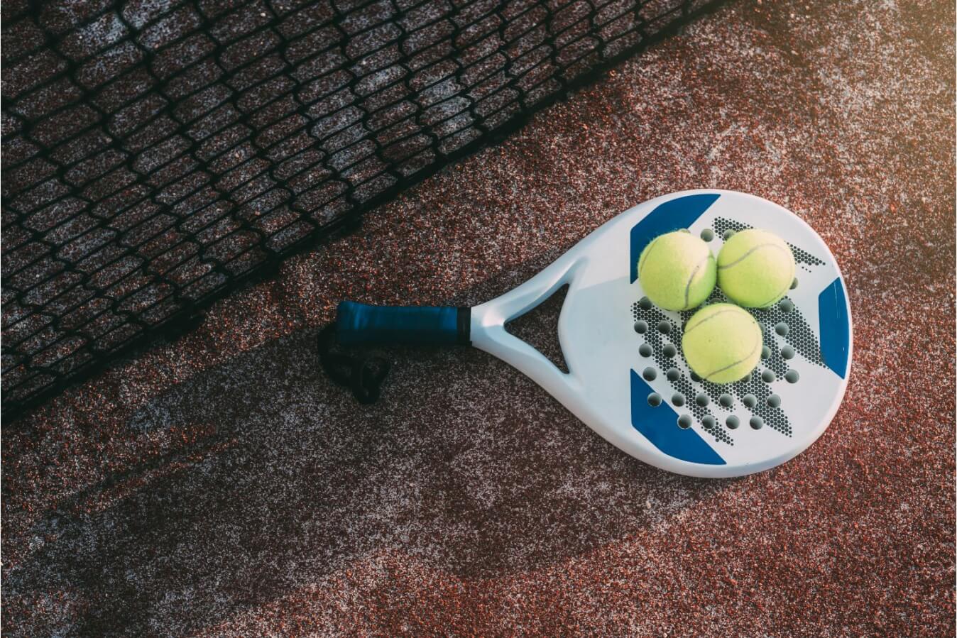 raquette de Padel de couleur blanc et bleu avec manche noir posée sur un terrain près du filet avec 3 balles jaunes sur le dessus