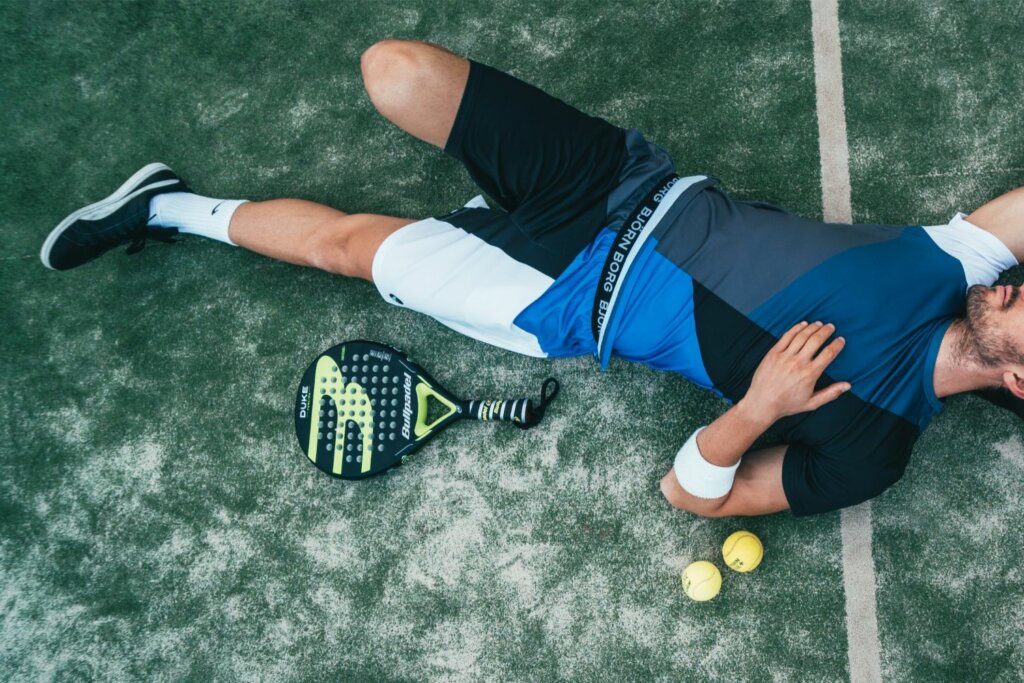 joueur de Padel avec chaussures noires allongé sur le sol d'un terrain près d'une raquette et 2 balles