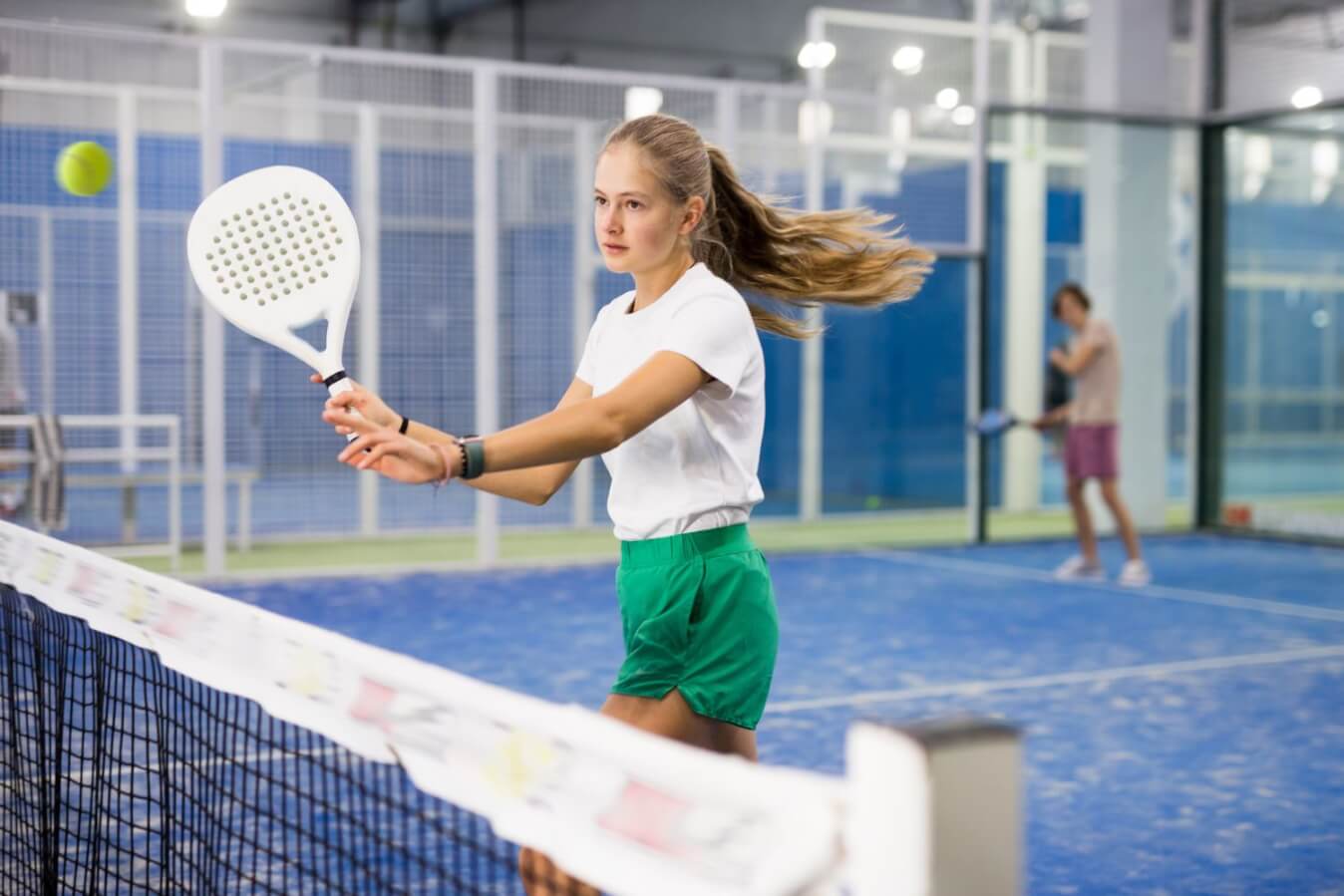 jeune joueuse de Padel en t-shirt blanc et short vert avec une raquette blanche à la main près du filet