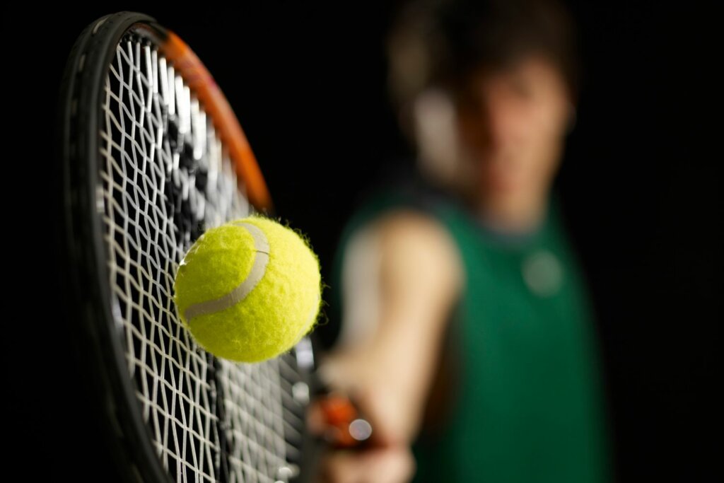au premier plan, les cordes d'une raquette de tennis entrant en contact avec la balle et un joueur en tenue vert
